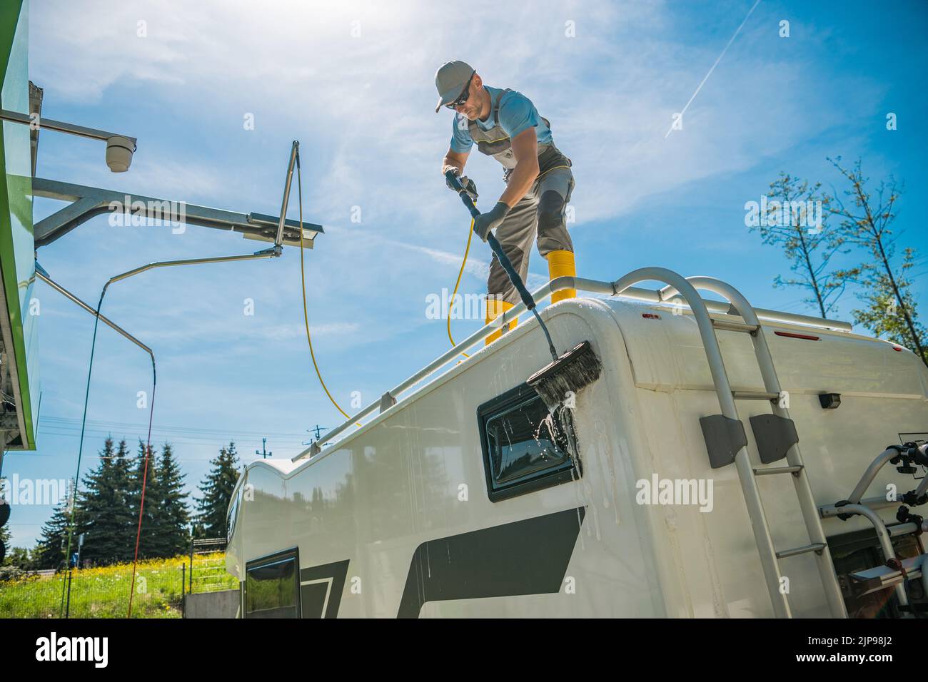 Caucasian Man Carefully Cleaning His White Recreational Vehicle with Car Wash Brush After the Trip . Summer Season. Blue Sky in the Background. Motorh Stock Photo