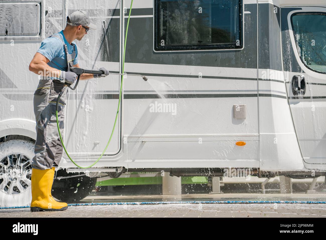 Caucasian Middle Aged Man in Rubber Boots Washing His White Camper Van with Pressure Washer at Self-Service Car Wash. Horizontal Photo. Motorhome Care Stock Photo