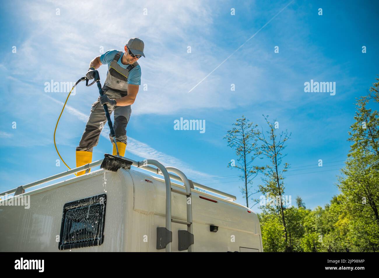 Caucasian Man in Uniform and Yellow Rubber Boots Standing on the Roof of White RV Cleaning the Surface with a Large Car Wash Brush. Clean Blue Sky and Stock Photo