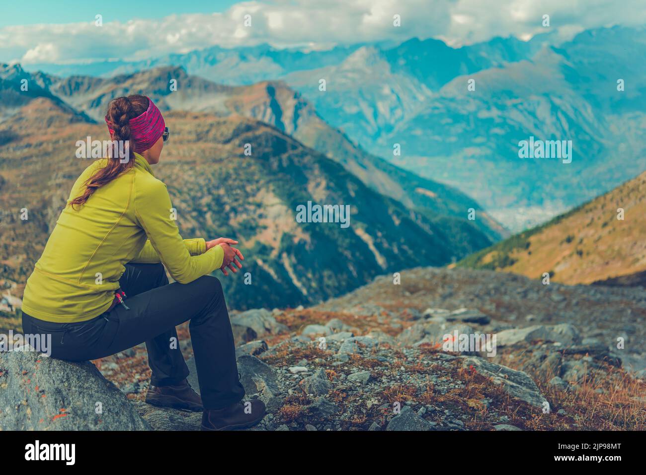 Caucasian Female Hiker Sitting Alone on a Stone on a Mountain Trail Relaxing and Enjoying Scenic Alpine Landscape in Front of Her. Travel and Adventur Stock Photo