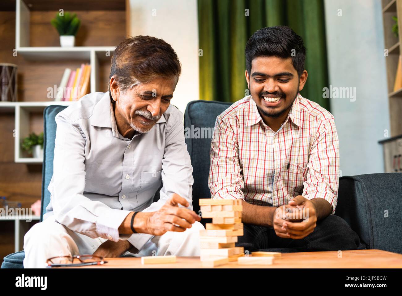 Excited grandfather with grandson playing tumbling tower game while sitting at home - concept of leisure activity, entertainment and family bonding. Stock Photo