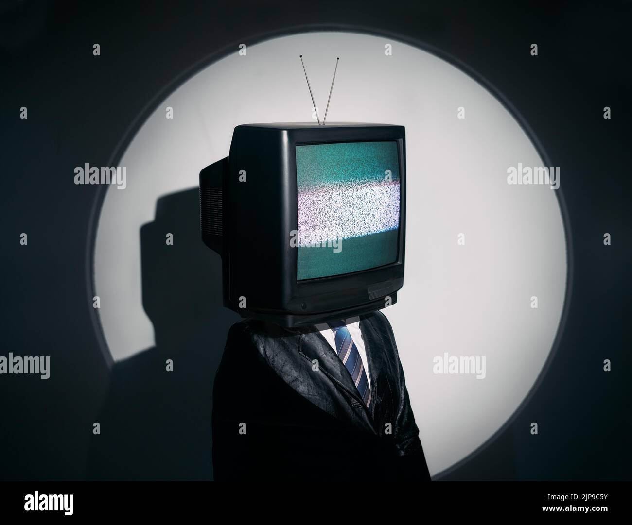 Man with TV instead of head. Media zombie concept with television addicted male in black coat with crossed arms posing on studio in the spotlight. Con Stock Photo
