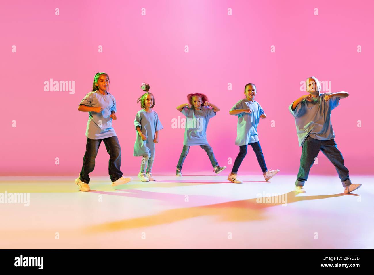 Hip-hop dance, street style. Happy children, little active girls in casual style clothes dancing isolated on orange background in purple neon light. Stock Photo