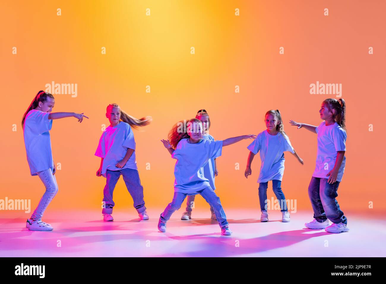 Group of children, little girls in sportive casual style clothes dancing in choreography class isolated on orange background in purple neon light. Stock Photo