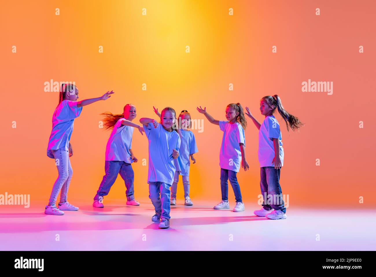 Hip-hop dance, street style. Happy children, little active girls in casual style clothes dancing isolated on orange background in purple neon light. Stock Photo