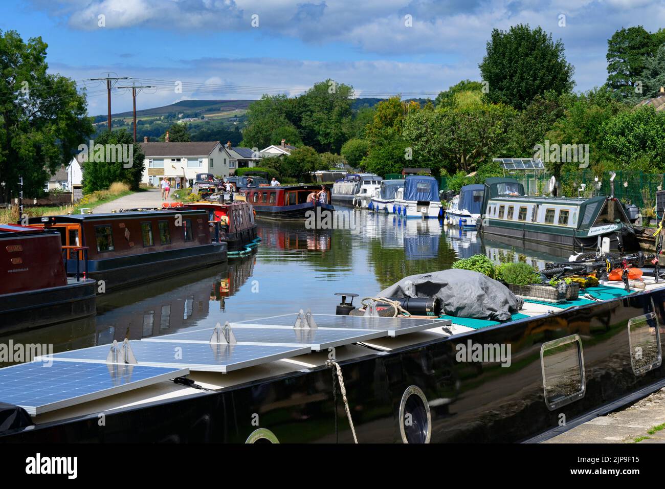 Barges at busy moorings, self-drive hire boat traveling, scenic sunny stretch of Leeds Liverpool Canal & towpath - Bingley, West Yorkshire England UK. Stock Photo