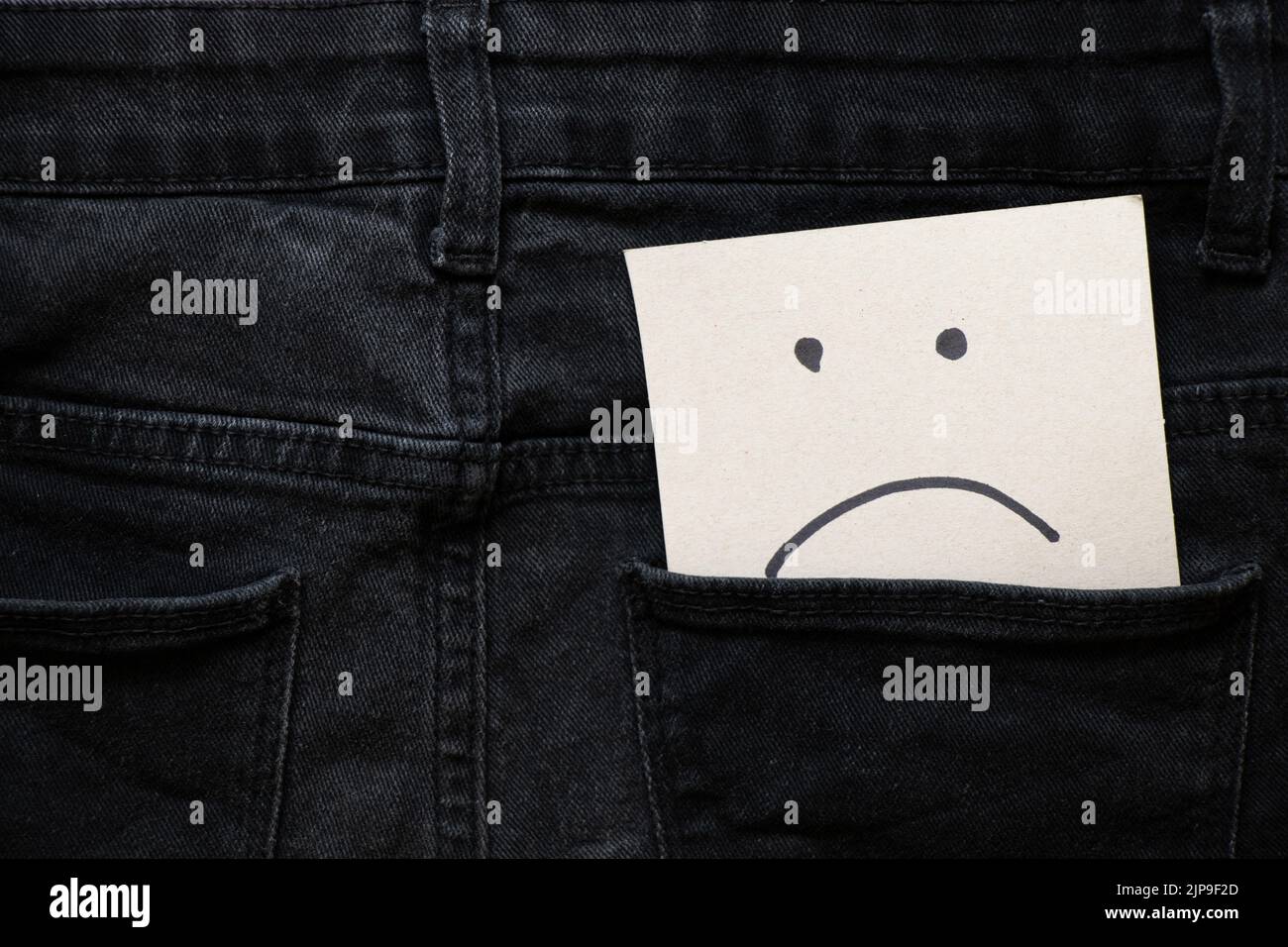 Sad emoticon drawn on paper lies in the pocket of gray jeans, bad mood, depression Stock Photo