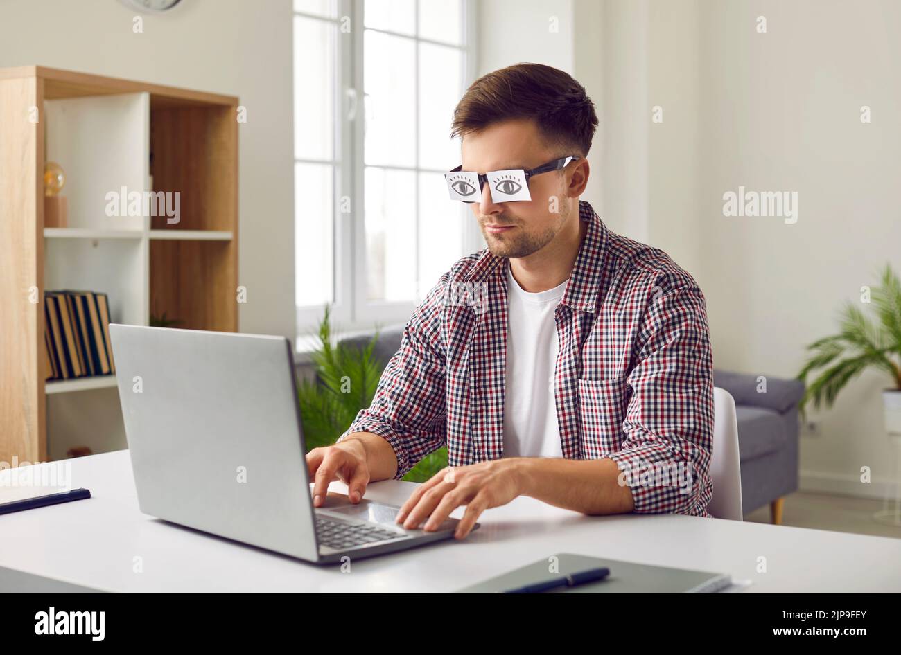 Tired sleepy man working on laptop and hiding his tiredness behind fake eye sticker glasses Stock Photo