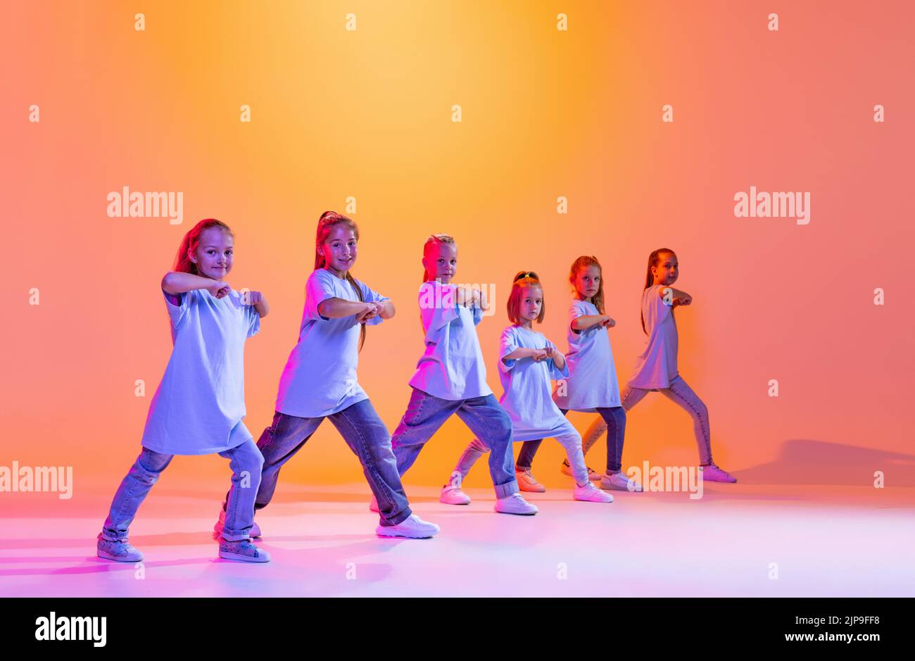 Portrait of cheerful, active little girls, happy kids dancing isolated on orange background in neon light. Concept of music, fashion, art, childhood Stock Photo