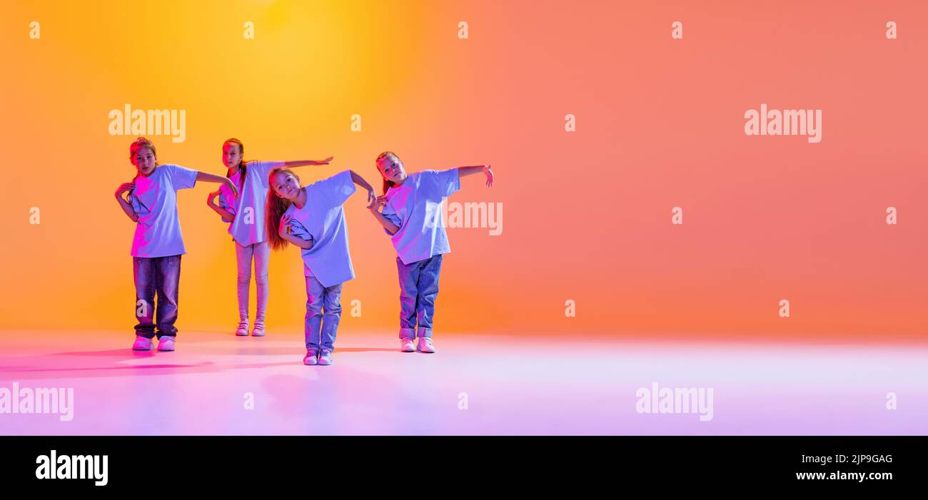 Dance group of happy, active little girls in t-shirts and jeans in action isolated on orange background in neon. Concept of music, fashion, art Stock Photo