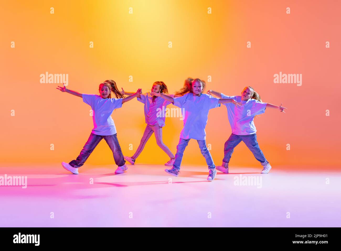 Group of children, little girls in sportive casual style clothes dancing in choreography class isolated on orange background in purple neon light. Stock Photo