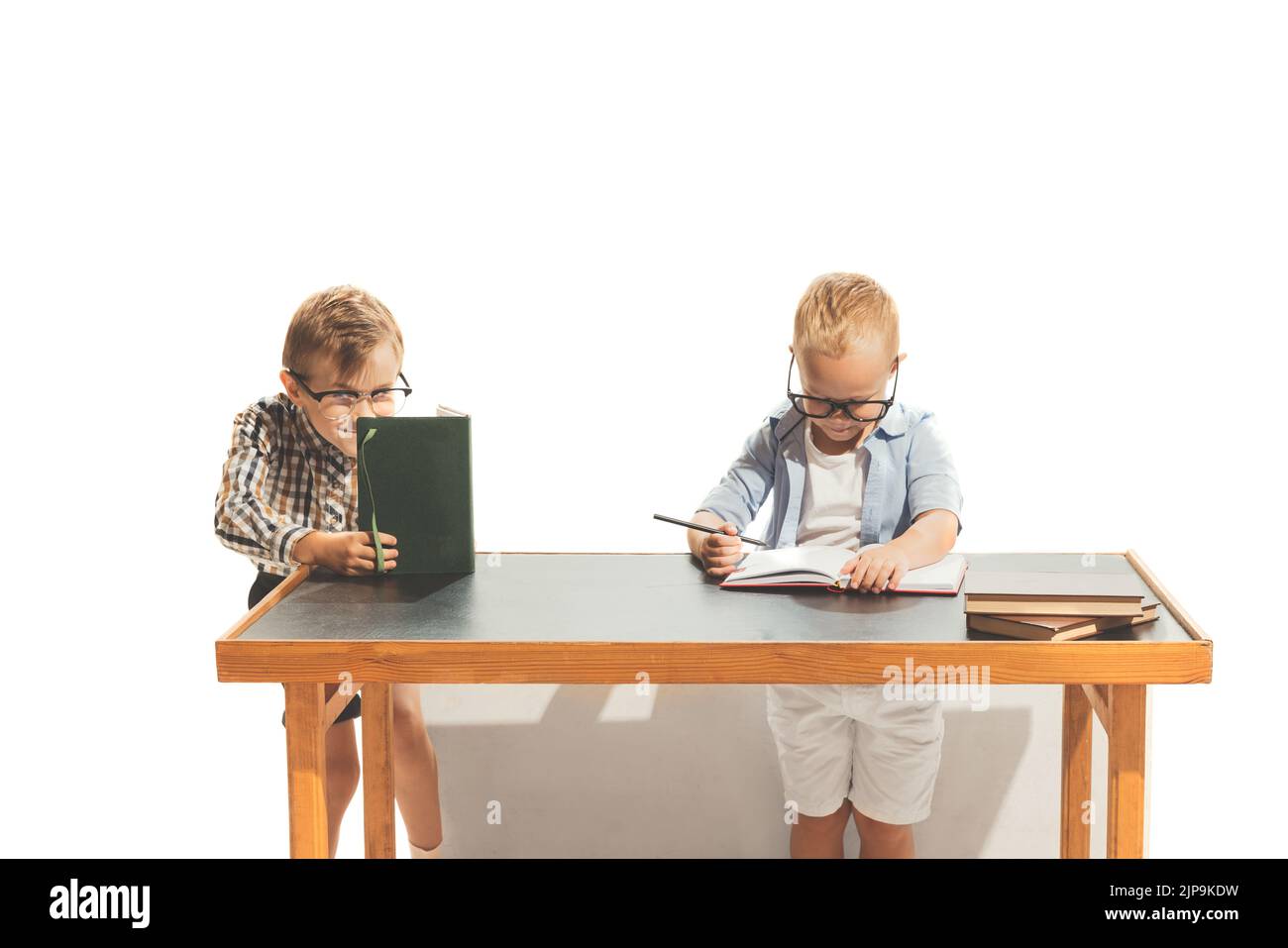 Portrait of two little boys, children, pupils sitting at desk, studying, doing homework isolated over white studio background Stock Photo