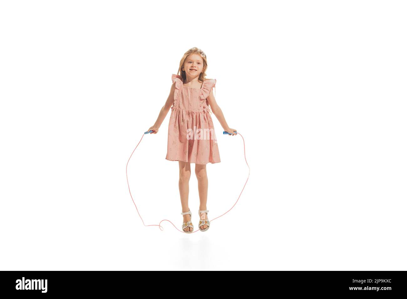 Portrait of beautiful little girl in stylish dress posing, jumping with rope, playing isolated over white studio background Stock Photo