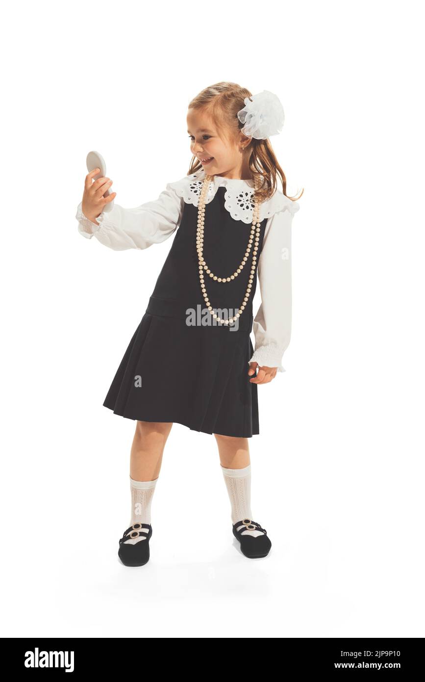 Portrait of little girl, child in school uniform with bow looking at mirror isolated over white studio background Stock Photo