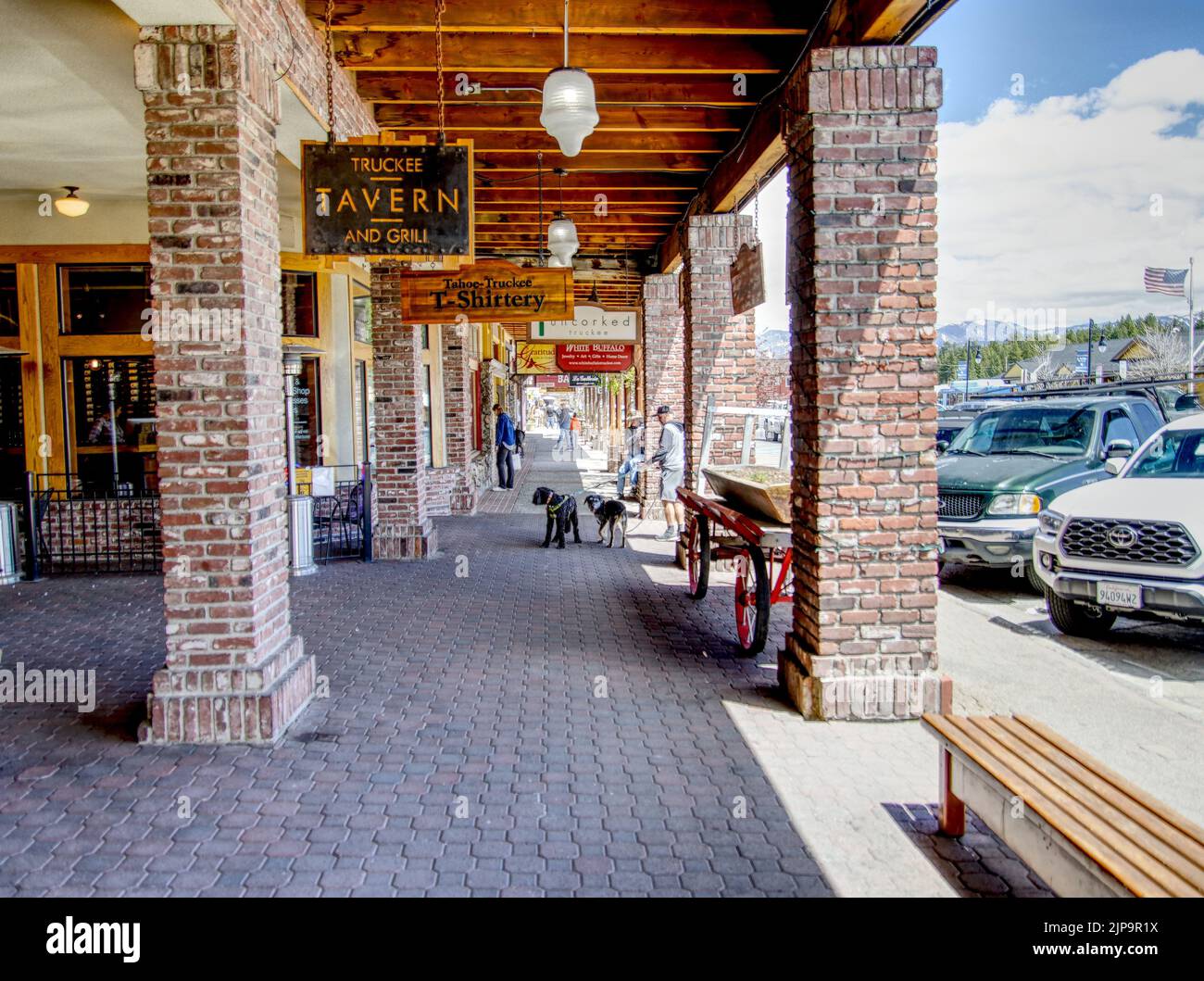 A crowdy street with brick columns, dogs, cars, benches, and a hanging tavern sign in Truckee, US Stock Photo