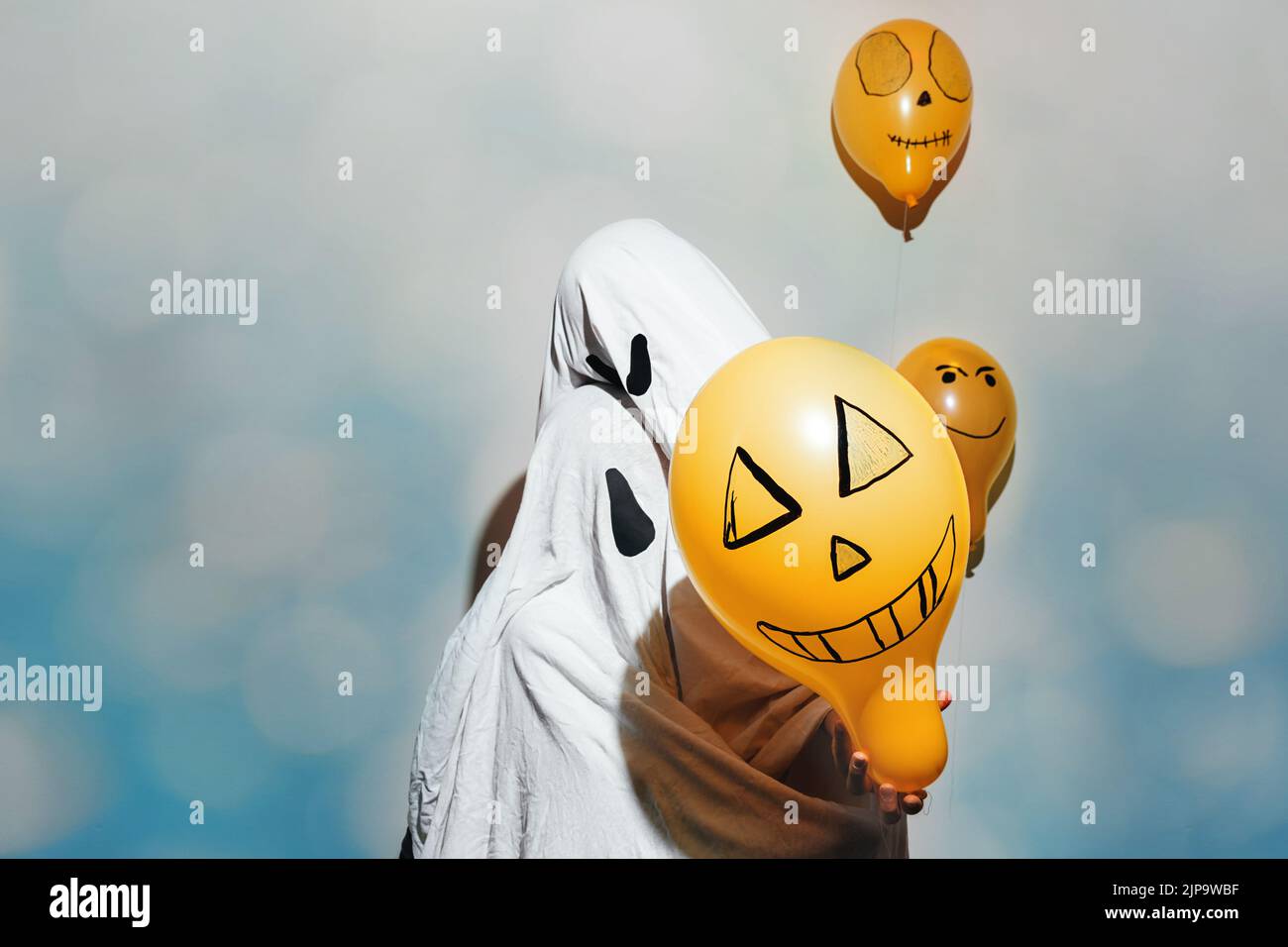 Happy Halloween. Ghosts hug. Children dressed in white costumes and painted orange balls on blue shimmering background. Different emotions of joy, anger. Festive decoration,concept of party at home. Stock Photo