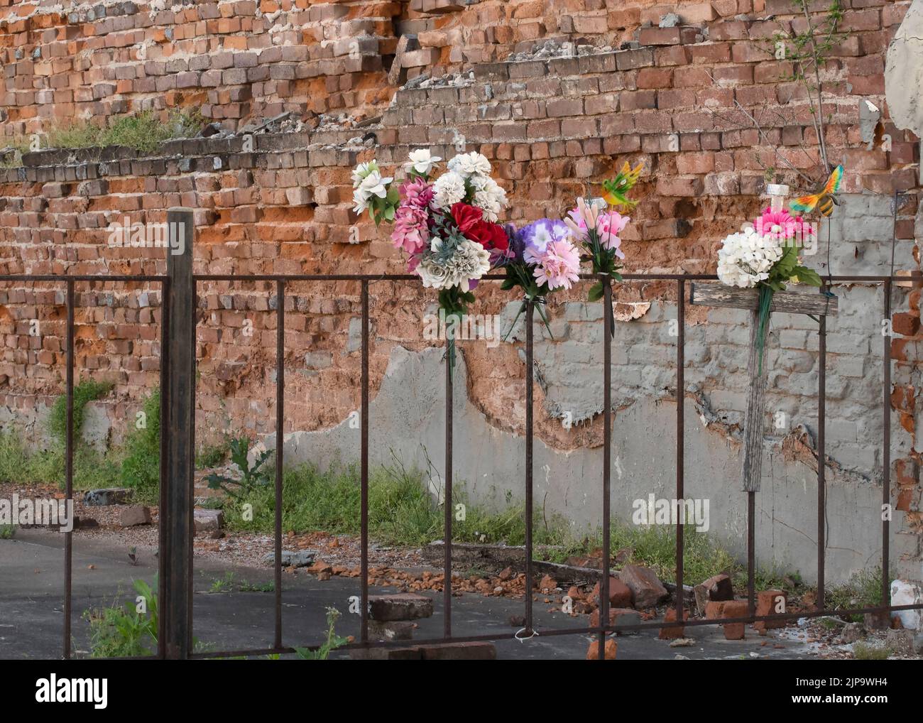 Memorial flowers on a iron fence with a wooden cross.  Old brick wall background. Stock Photo