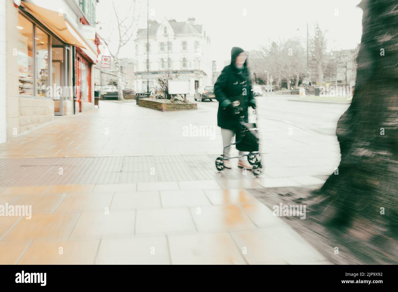 Senior woman walking with a shopping walking frame down the high street in Ilkley town centre on a rainy winter day (ICM) West Yorkshire, England, UK Stock Photo