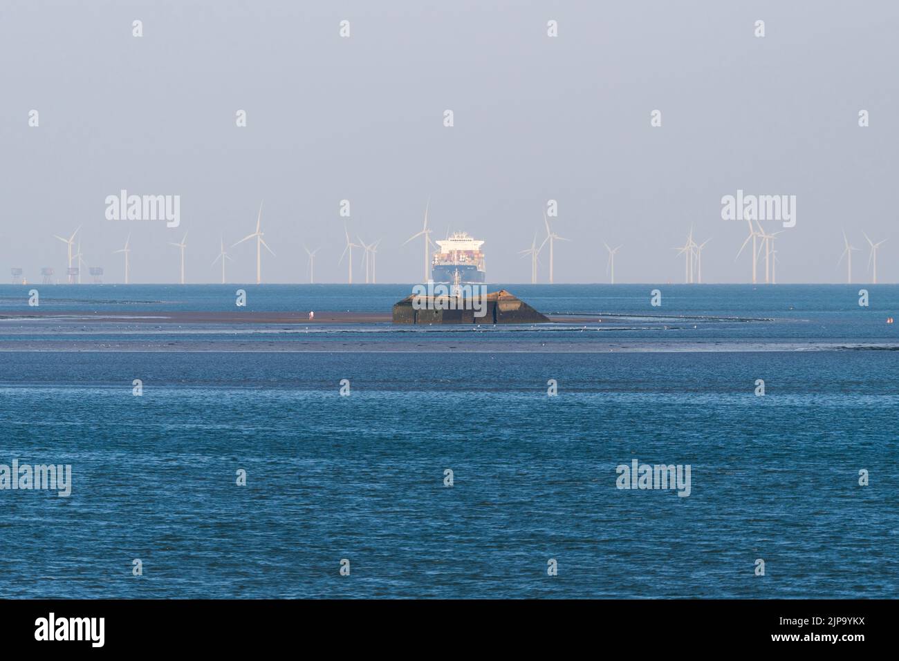 Distant cargo ship navigating through offshore windfarms in Thames Estuary, with walkers far out from shore at Mulberry Harbour section off Southend Stock Photo