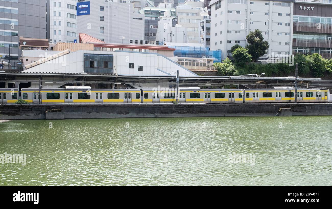 The JR Ichigaya Train Station is located along a river with the yellow Toei Subway-Shinjuku Line train on a cloudy summer day. Stock Photo
