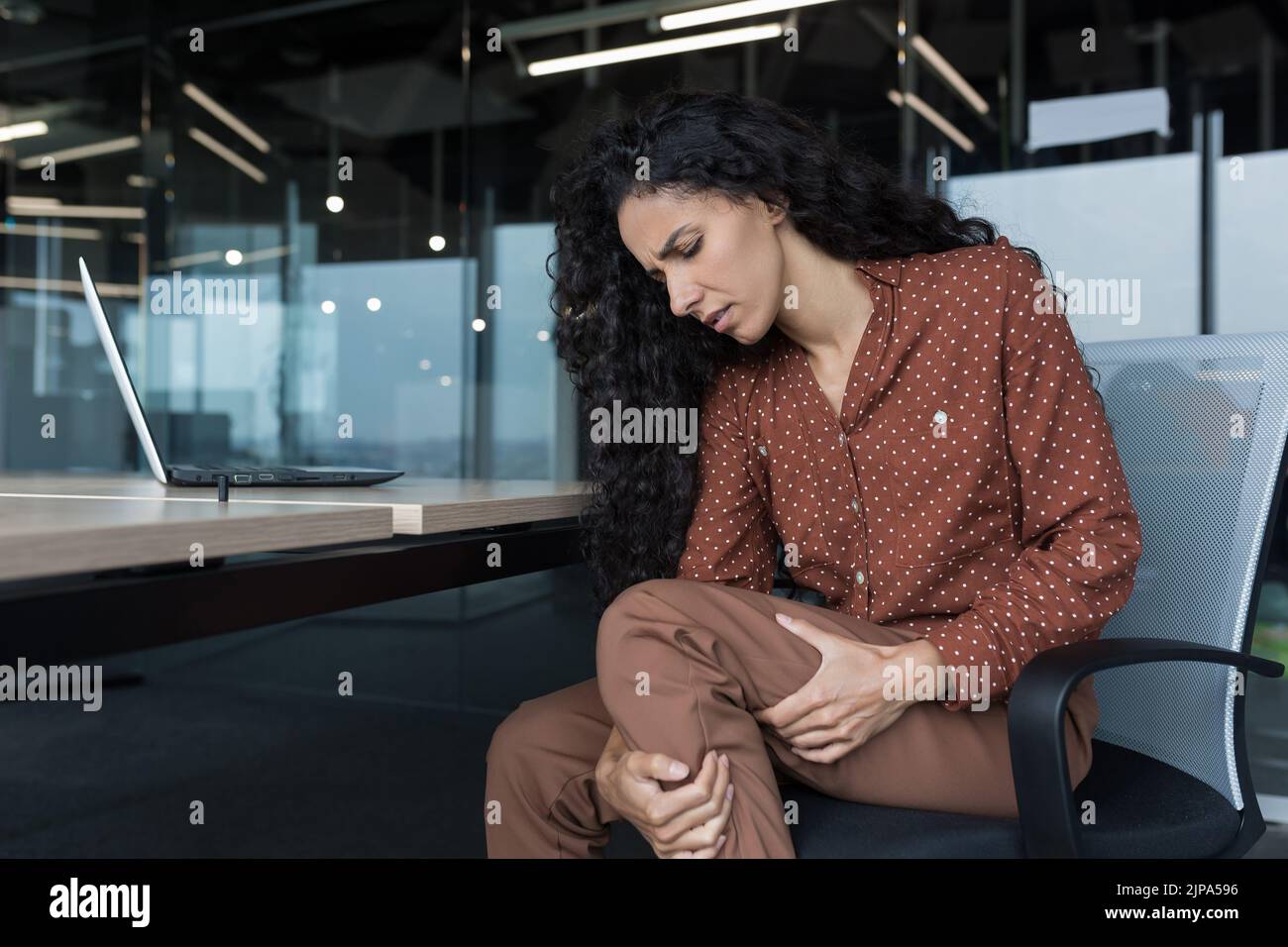 Young beautiful arab woman working in modern office with laptop, curly business woman has severe pain in leg muscles, varicose veins, massaging her leg Stock Photo