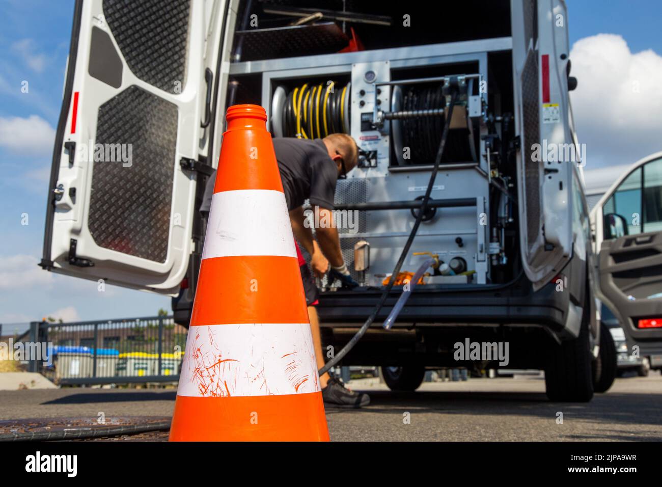 Mitarbeiter eines Kanalreinigungsunternehmens bei der Reinigung eines Straßenablaufs. Im Vordergrund das Baustellenhütchen, im Hintergrund das Einsatz Stock Photo