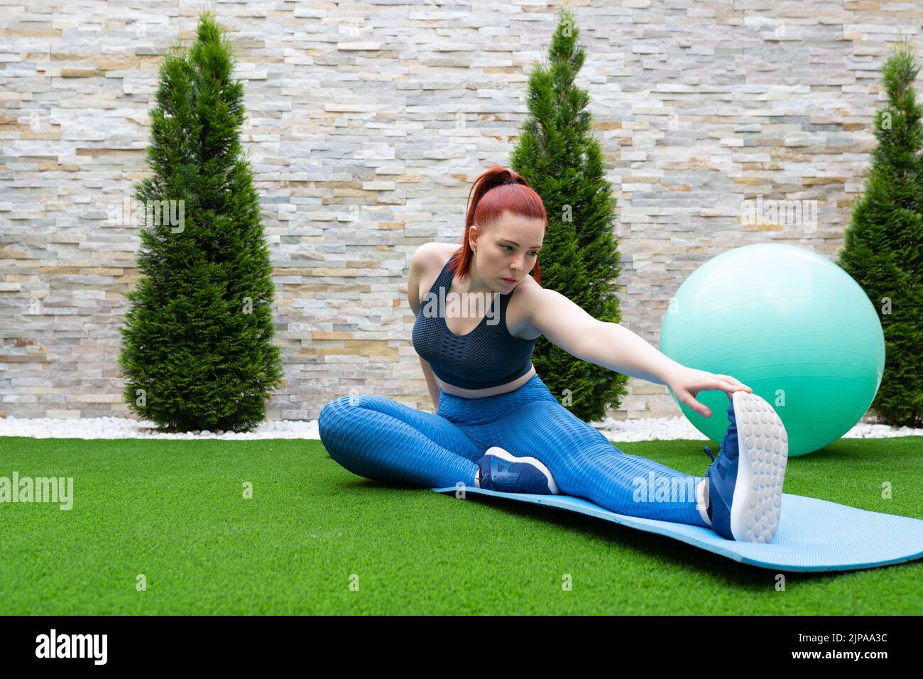 young woman with red hair, practising leg stretching exercises on a mat in the garden of her house. Sporty girl training. concept of health and well Stock Photo
