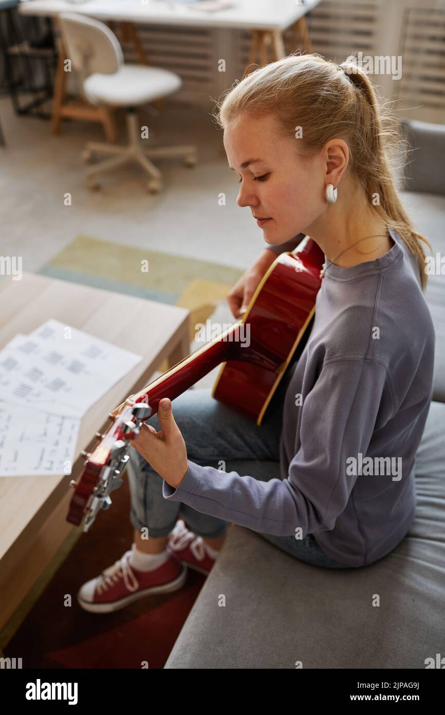 Vertical portrait of young woman playing guitar at home while sitting on sofa and composing music Stock Photo