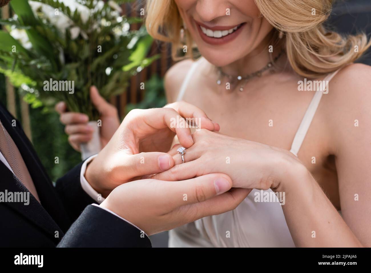 Cropped view of groom wearing ring on finger of smiling bride with blurred bouquet Stock Photo