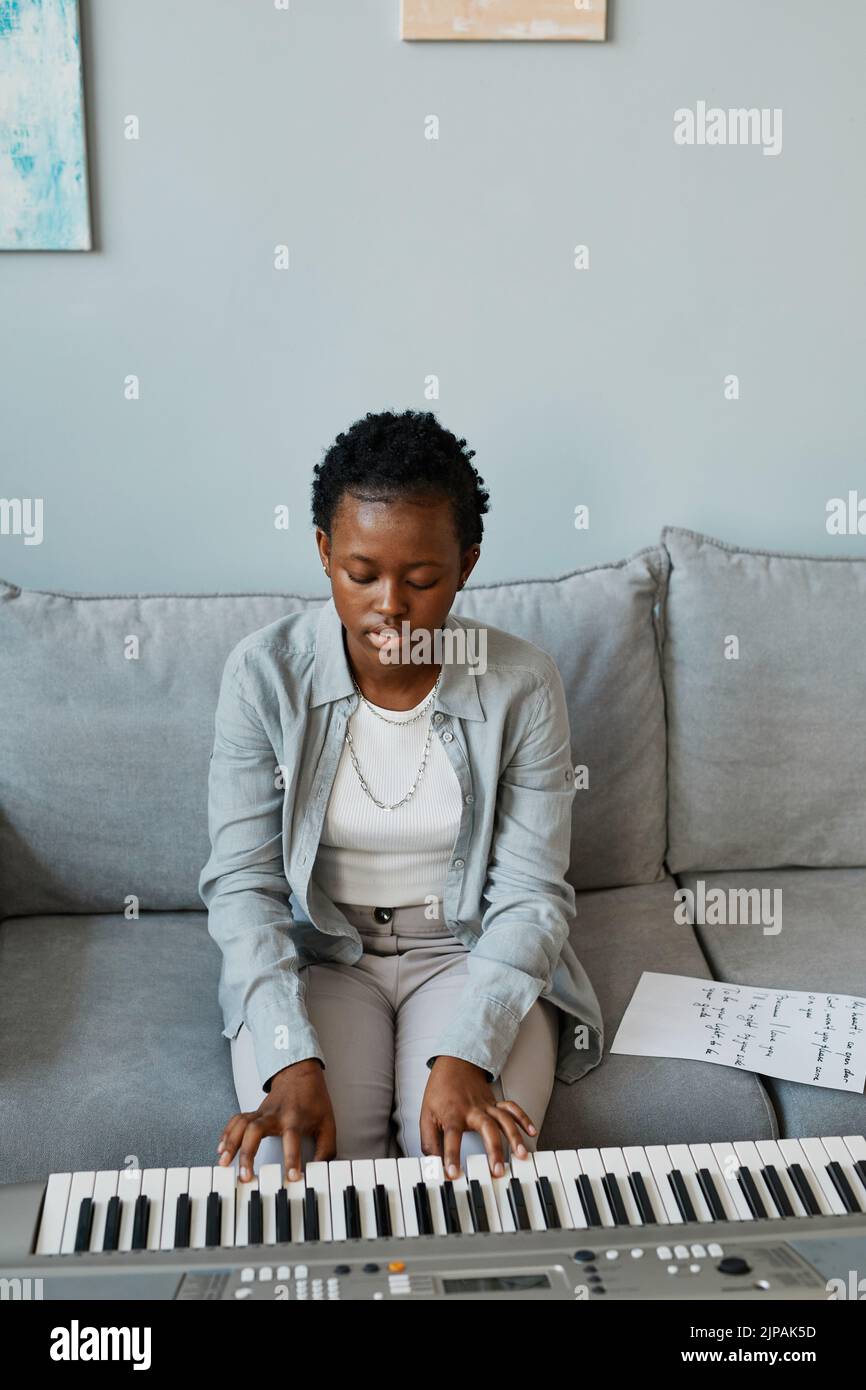 Vertical portrait of young black woman playing synthesizer at home and composing music in minimal home setting, copy space Stock Photo