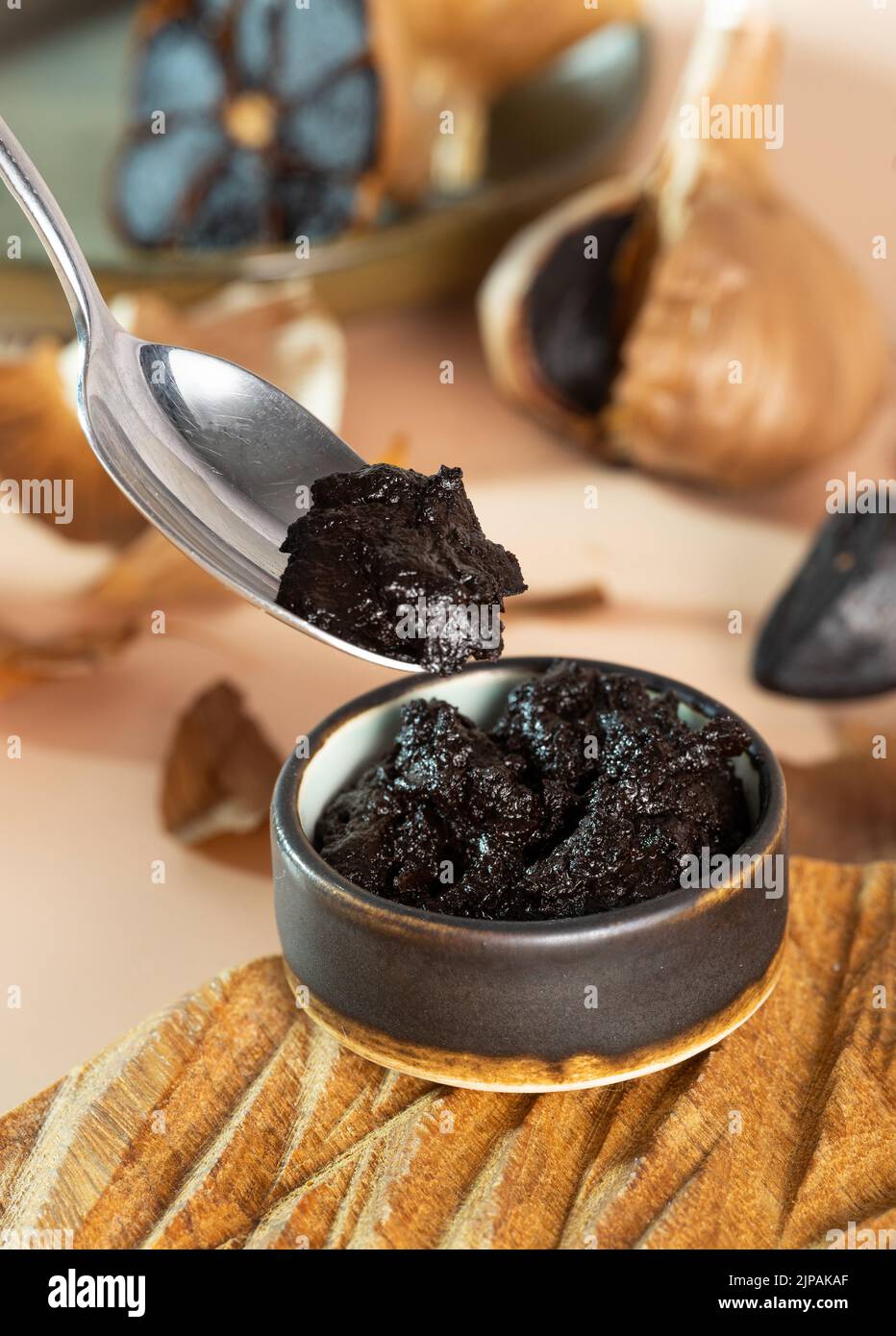 A small clay bowl with black garlic paste on a handmade wooden plate. Spoon with black aioli. Fermented garlic bulb in the background. Healthy Stock Photo