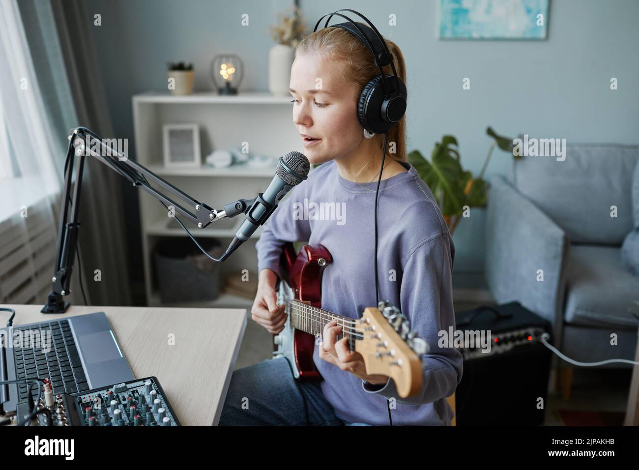 Side view portrait of blonde young woman playing electric guitar and singing to microphone in home recording studio, copy space Stock Photo