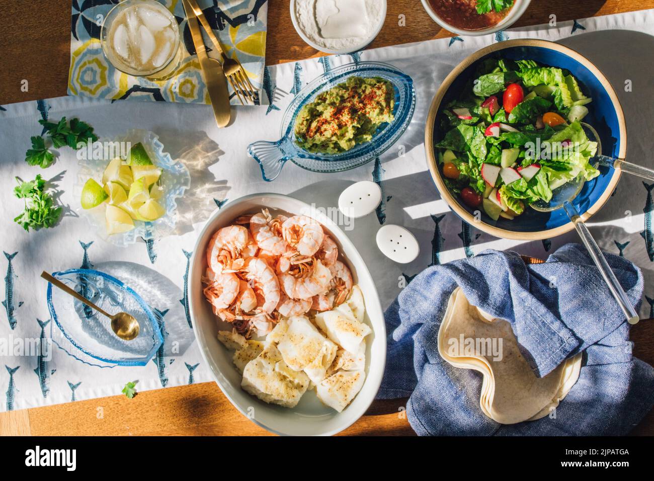 summer Seafood supper table spread to make fish tacos, prawns, guacamole, lime, tortilla, salsa on blue and white fish tablecloth in the sun Stock Photo