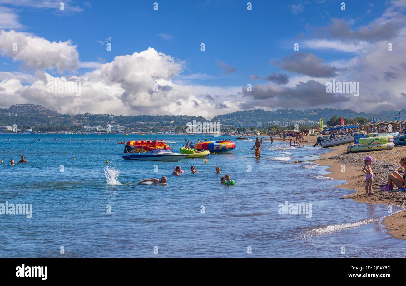 eautiful view of people on beach on sunny summer day. Blue sea water on front and mountains covered with white clouds on blue sky background. Rhodes. Stock Photo