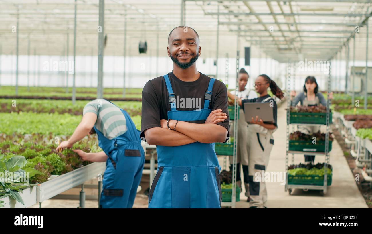 Smiling african american man posing with arms crossed while farm workers using laptop manage