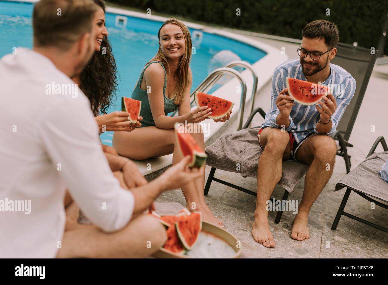 Group of young people sitting by the swimming pool and eating watermelon in the house backyard Stock Photo