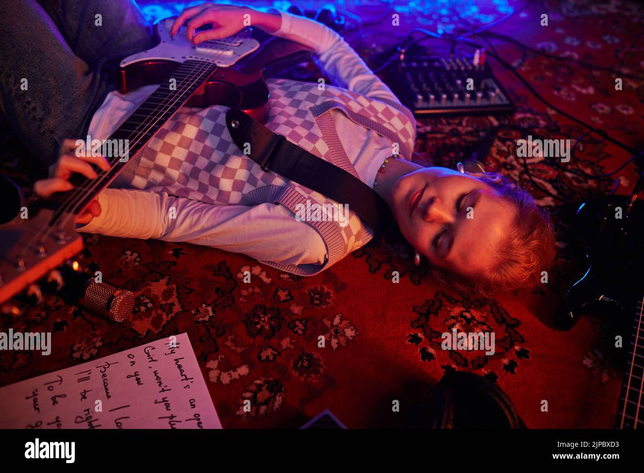 Closeup of young woman playing guitar while lying on carpet lit by dim red lights Stock Photo