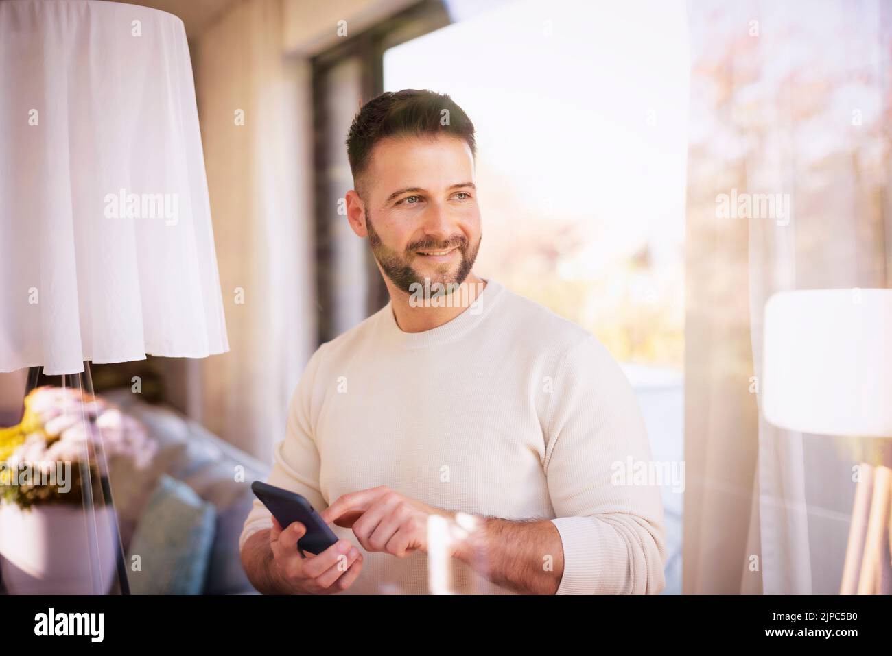 Shot of handsome man wearing casual clothes and text messaging while standing at the window at home. Stock Photo