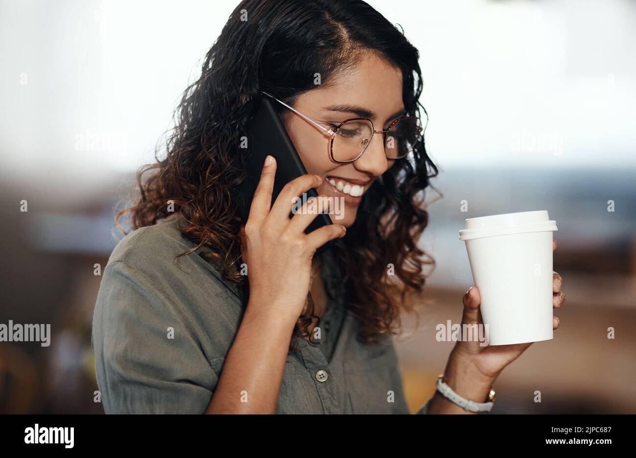 Young woman talking on her phone, drinking coffee, and smiling in cafe shop. Lady wearing glasses, using technology to network and connect while Stock Photo