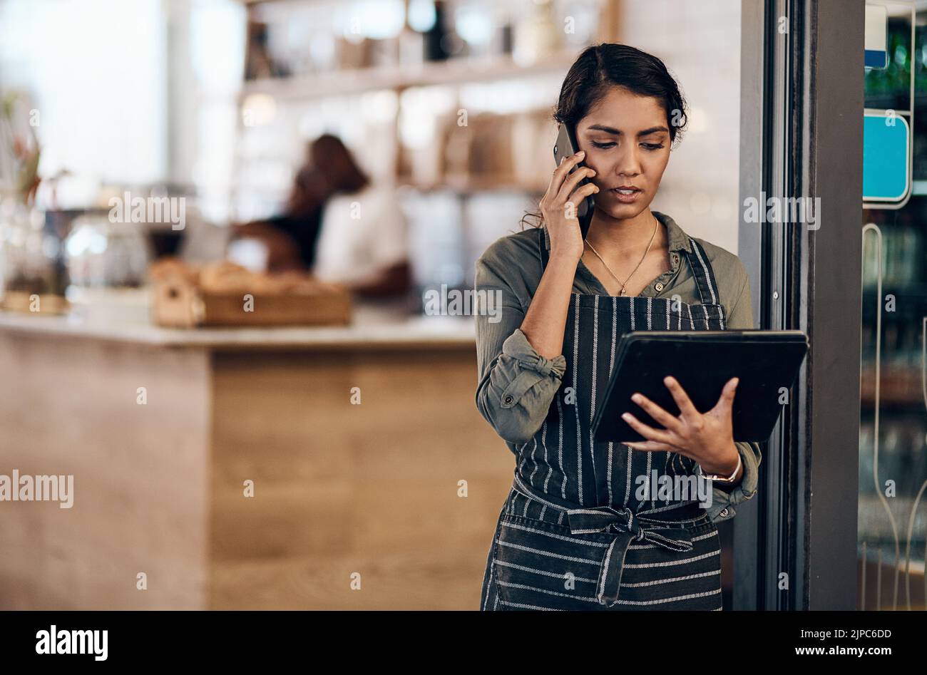 Female cafe business owner using phone talking, ordering and reading on tablet in her store. Serious businesswoman, entrepreneur or employee standing Stock Photo