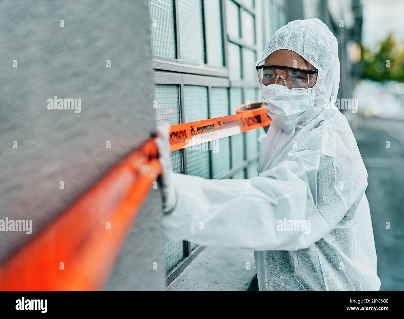 Covid healthcare worker responding to a biohazard in a public area using barrier tape outside. First responder in protection hazmat suit and mask Stock Photo