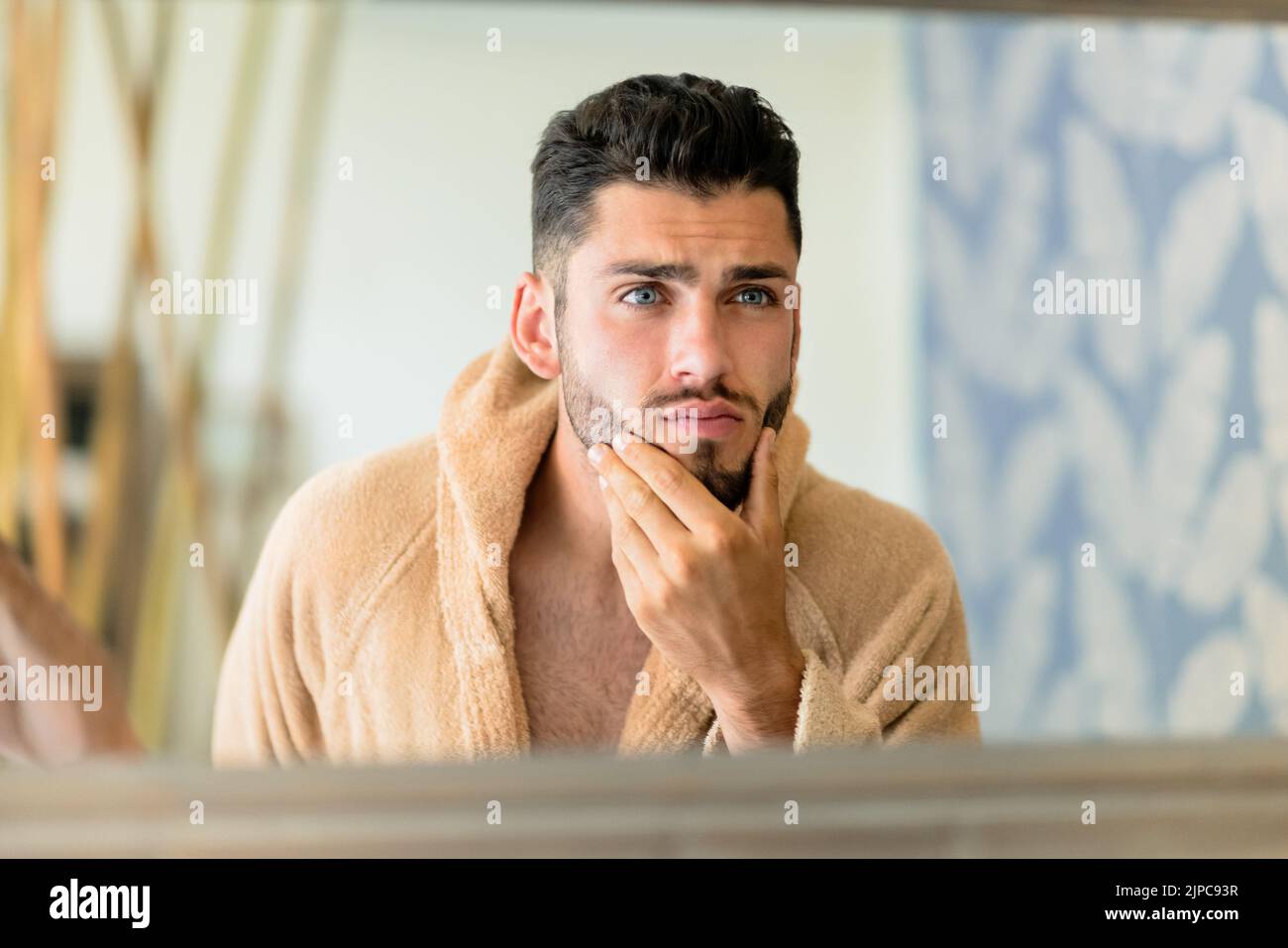 Young male in warm bathrobe touching beard and looking in mirror during