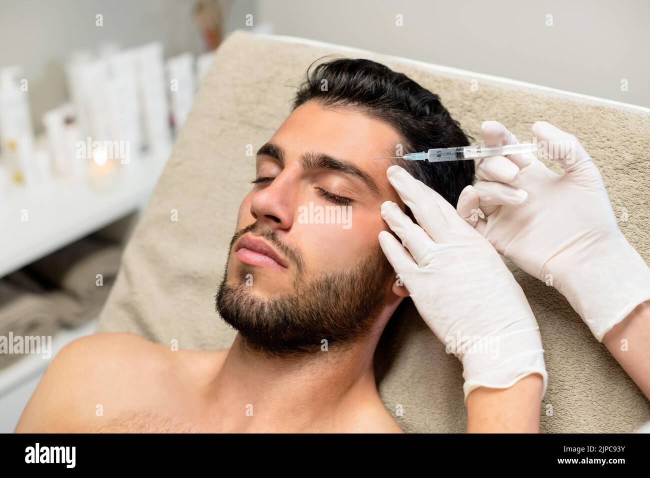 From above anonymous beautician in latex gloves injecting botulinum toxin into forehead of bearded young male client during cosmetology session in bea Stock Photo