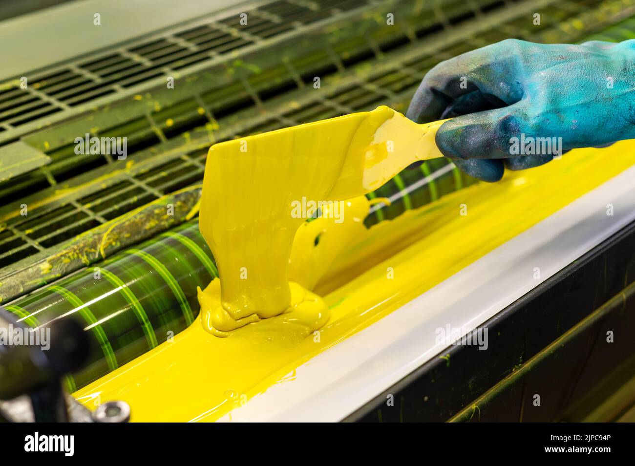 From above anonymous employee in blue rubber glove using spatula to smear yellow paint on rollers of industrial offset printer Stock Photo