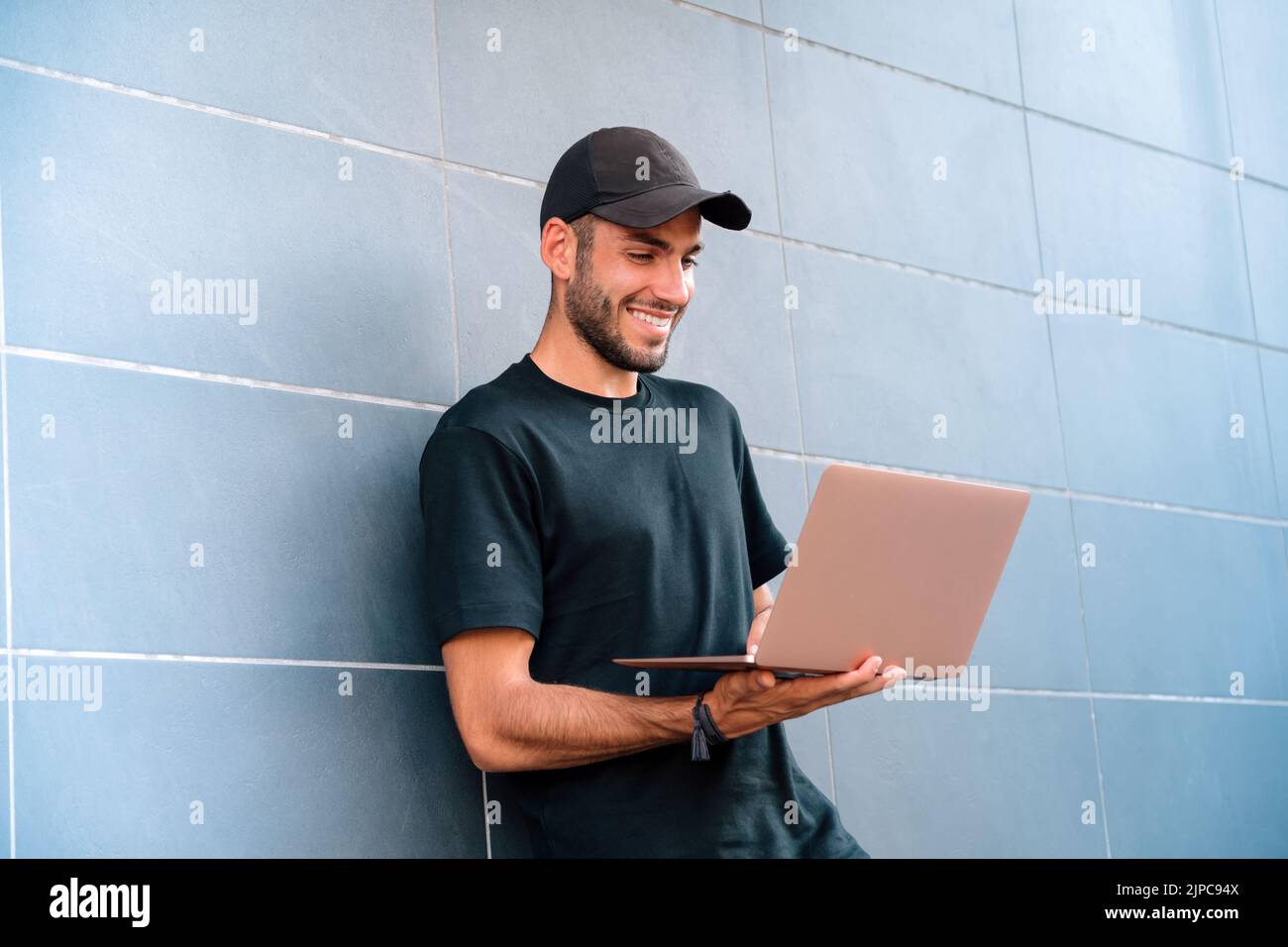 Low angle of cheerful young man in black t shirt and cap smiling and browsing netbook while leaning on tiled wall of modern building on city street Stock Photo