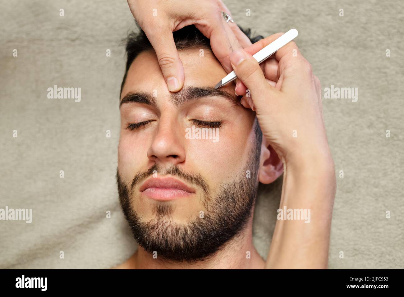 Top view of anonymous female cosmetician using tweezers to pluck eyebrow hair of calm young male client during work in beauty studio Stock Photo