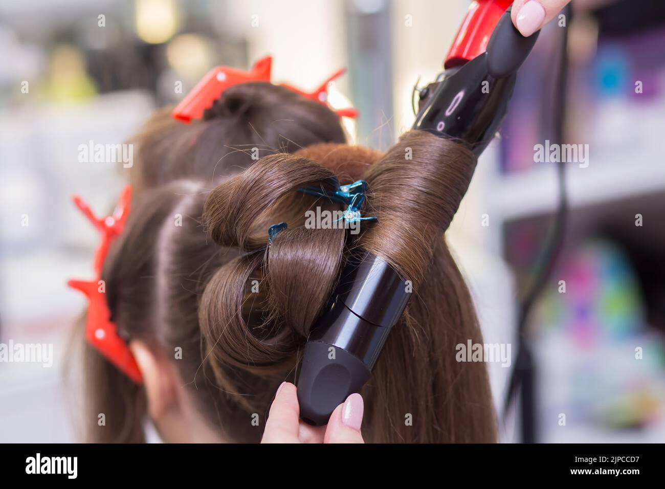 Close-up hot tongs curl a strand of hair on a woman's head. The hairdresser makes a hairstyle for a young woman. Barber shop, business concept. Beauty salon, hair care. Stock Photo