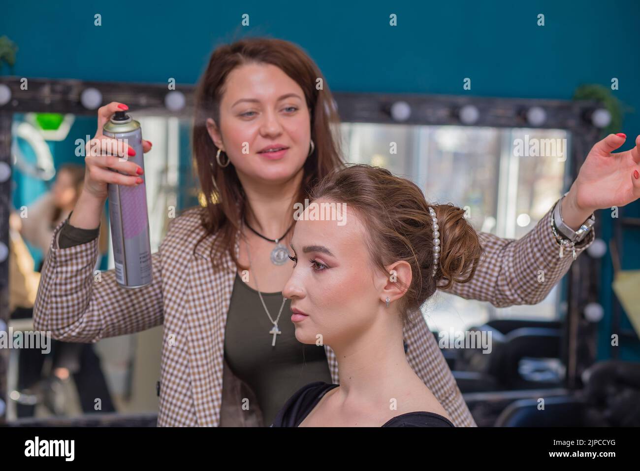 A young woman master with a smile sprinkles varnish on the client's hair. The hairdresser makes a hairstyle for a young woman. Barber shop, business concept. Beauty salon, hair care. Stock Photo