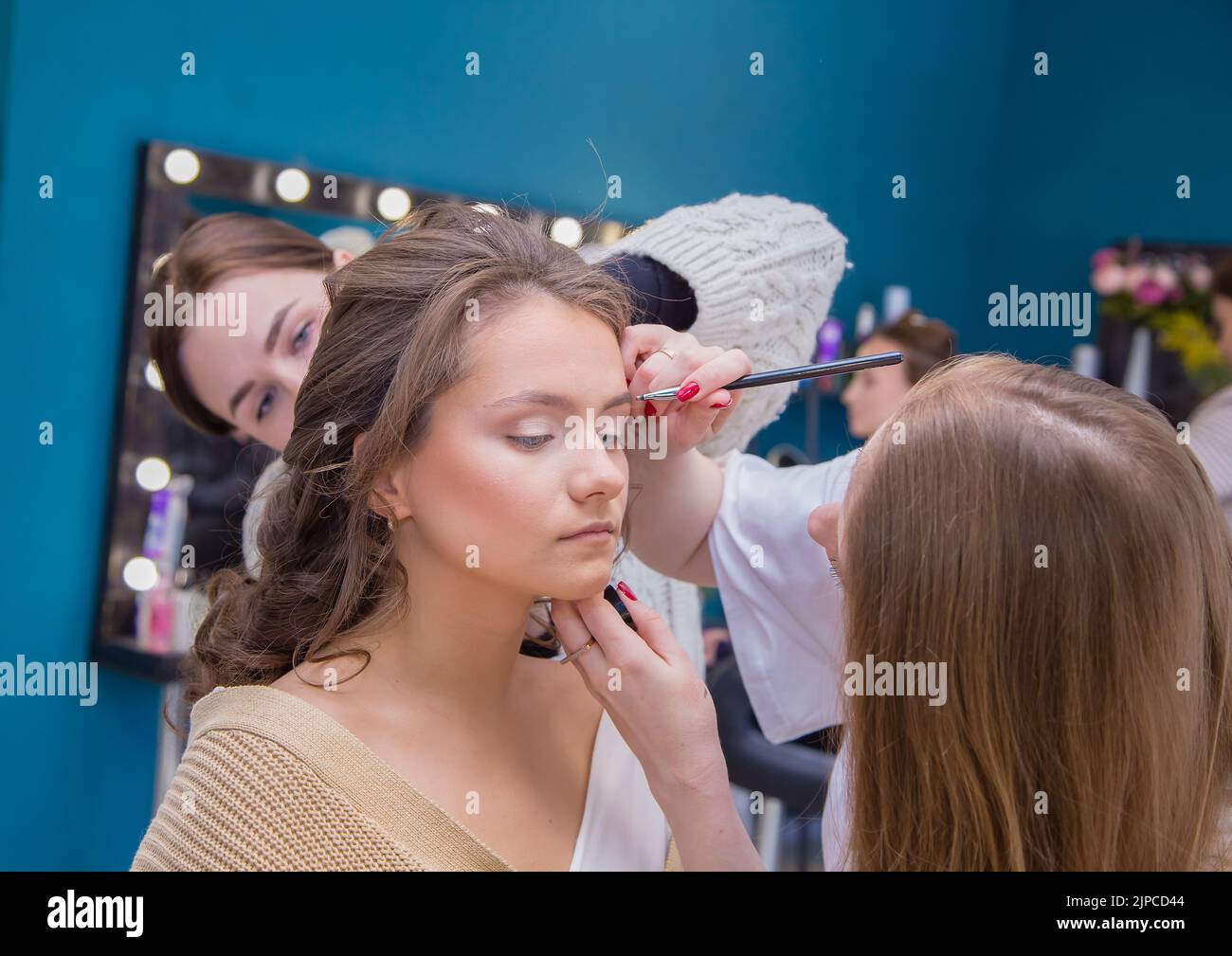 Two masters carefully do makeup and hair for a girl in the salon. Make-up artist and hairdresser create an image of a young woman. Business concept - beauty salon, facial skin and hair care. Stock Photo