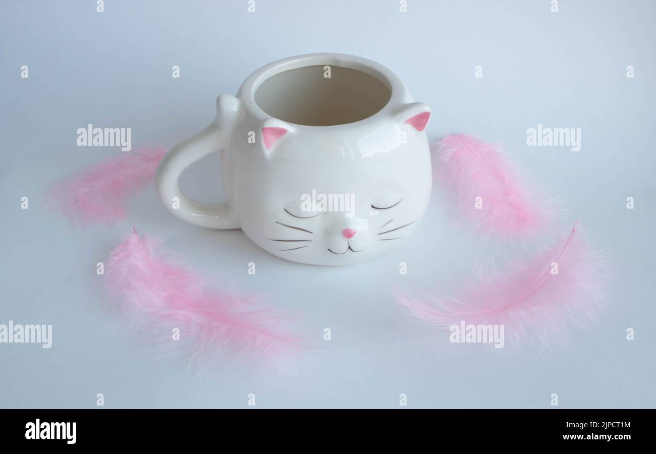 White coffee mug in the shape of a cat with pink ears, close-up on a white background with pink feathers. Stock Photo
